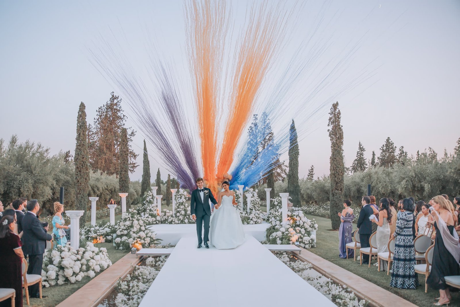 Western ceremony aisle among olive trees with plumes of coloured smoke
