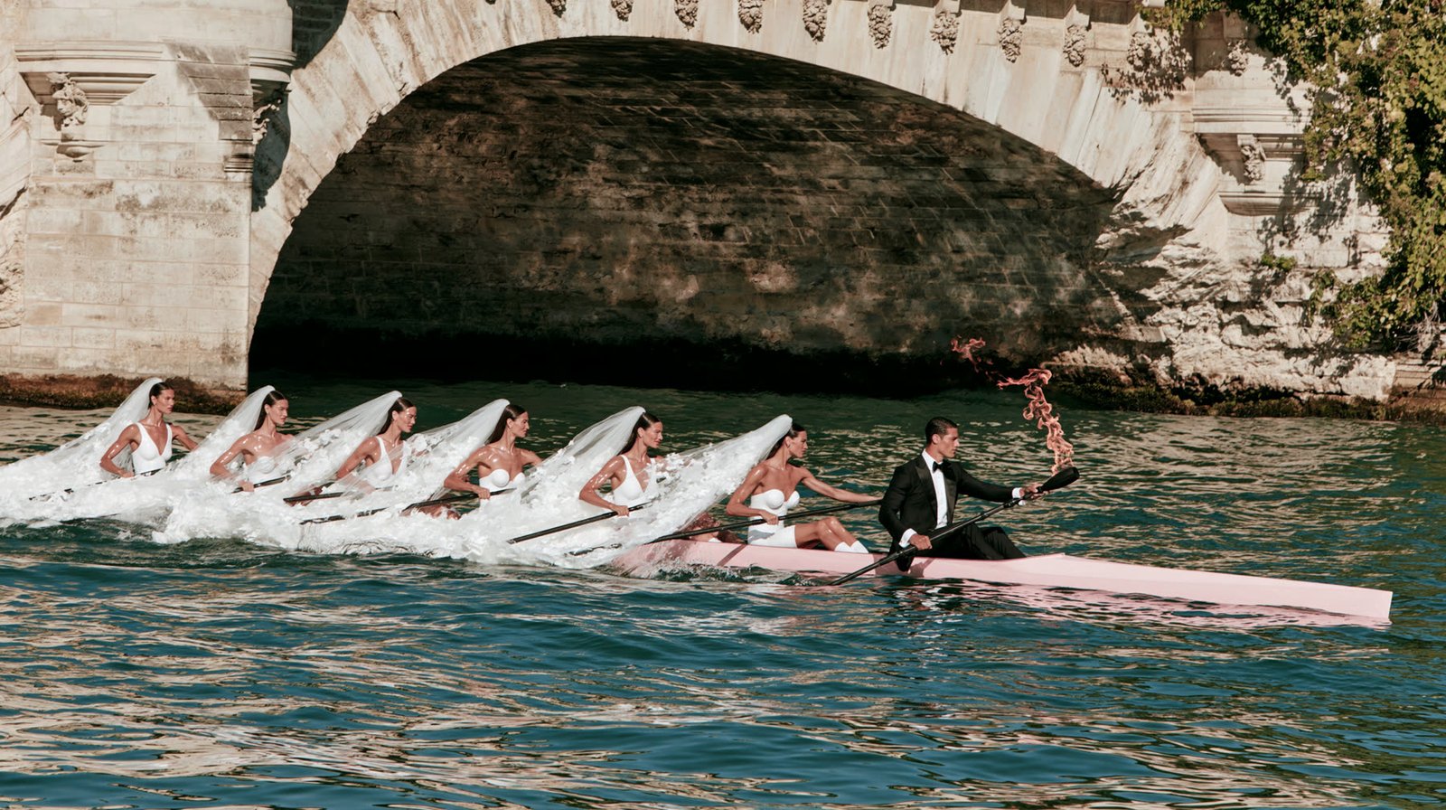 Brides rowing a white boat past a stone bridge on the Seine, echoing the Paris 2024 opening ceremony